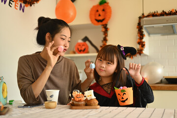 Cheerful little girl and mother decorating Halloween cupcakes with different monsters at kitchen table