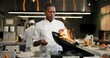 © CineLens2024/peopleimages.com - Black man, chef and fire with frying pan in kitchen at restaurant for fine dining, meal and creativity. Employee, professional and cook with food for culinary, hospitality and catering service