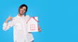 © Iryna - Smiling Woman Holding a Small House Model Against a Blue Background with Copy Space