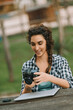 © Giulio_Fornasar - woman with curly braided hair and a checkered shirt sits at an outdoor table, smiling and examining her camera. The presence of a laptop suggests she is reviewing her photos