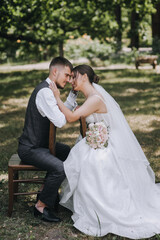 Wall Mural - A stylish groom in a gray suit and a bride in a white dress with a bouquet are sitting hugging a chair outdoors in a park on green grass. Wedding photography, portrait of stylish newlyweds.