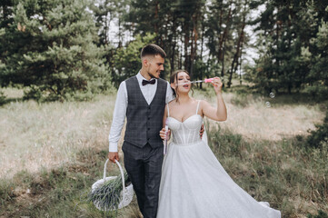 Wall Mural - A stylish fashionable groom and a beautiful young bride in a white dress are walking in the forest with a basket and soap bubbles in their hands. Wedding photo of happy newlyweds.