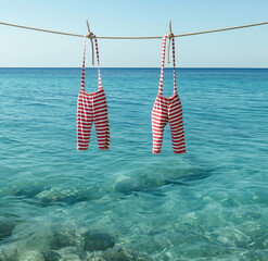 Wall Mural - Two red and white striped swimsuits hang on a clothesline above a clear blue sea. Minimal beach vacation concept.