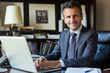 © forenna - Professional portrait of a successful male businessman seated at his desk, using a laptop and smiling confidently at the camera, perfect for illustrating financial expertise and co