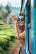© BetterPhoto - A cheerful young woman in sunglasses leans out of a train window, waving and enjoying the scenic green landscape.