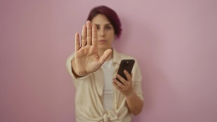 Canvas Print - Young caucasian woman holding smartphone with confident and serious expression using stop hand sign gesture for defense, isolated over pink background