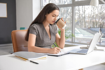 Wall Mural - Pretty businesswoman writing on clipboard and drinking coffee at table in light office