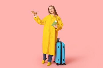 Sticker - Smiling young woman in raincoat with wooden airplane and passport on pink background. Travel concept