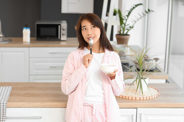 Wall Mural - Pretty young Asian woman in pajamas with tasty yogurt at home