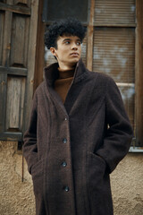 Wall Mural - Fashionable young man in brown coat and turtleneck sweater standing in front of old building with window in background