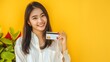 © The Little Hut - Portrait young beautiful asian woman enterpriser happy smile wearing white shirt and red plants holding credit card isolation on yellow background Money online payment concept : Generative AI