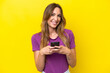© luismolinero - Young caucasian woman isolated on yellow background looking at the camera and smiling while using the mobile