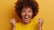© Pinklife - An excited woman with a curly afro and a bright smile raises her fists in the air. She stands against a plain yellow background, radiating happiness and enthusiasm.