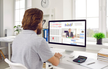 Young man working on office computer. Businessman or financial accountant sitting at desk, looking at screen of modern desktop PC, and using lots of different business apps. View over shoulder