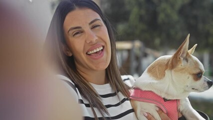 Wall Mural - A young, beautiful hispanic woman in an urban park is taking a selfie with her chihuahua dog, smiling happily in the sunlight while enjoying the outdoors.