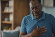 © Dinusha - a black american man in a blue shirt having chest pain, holding his hands on his heart area and standing near a sofa at home. suffering from chest pain or angina. close-up, pained expression.