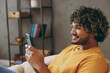 © ViDi Studio - Close up young happy Indian man wear orange casual clothes hold use mobile cell phone sits in armchair stay at home hotel flat rest relax spend free spare time in living room indoor. Lounge concept.