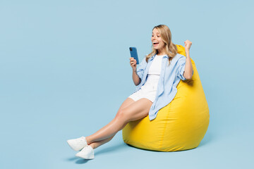 Wall Mural - Full body young happy woman wears white top shirt casual clothes sit in bag chair hold in hand use mobile cell phone do winner gesture isolated on plain light blue cyan background. Lifestyle concept.