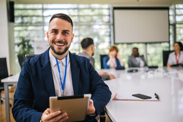 Portrait of young businessman sitting in his office in front of his colleagues with tablet in his hands.