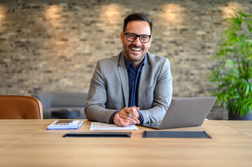 Wall Mural - Portrait of handsome smiling entrepreneur with laptop and documents on desk sitting in modern office