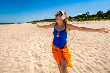 Summer holidays on beach with music. Beautiful young woman in blue swimsuit and orange sarong listening to music in headphones and dancing on white sandy beach. Front view