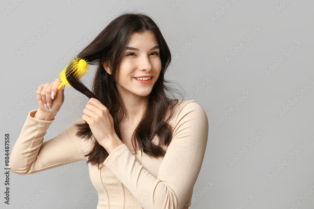 Beautiful young woman brushing her hair on grey background