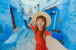 © Davide Angelini - Young woman with red dress visiting the blue city Chefchaouen, Marocco - Happy tourist taking selfie picture in Moroccan city street - Travel and vacation lifestyle concept
