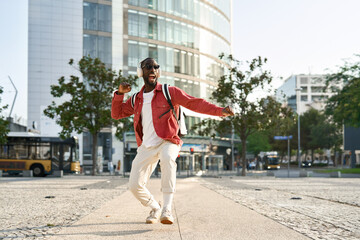 Happy cool young African Black man student hipster wearing headphones, sunglasses and backpack feeling joy having fun listening music and dancing on city street outdoors on sunny day, full body.