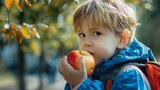 A young child enjoying a healthy snack outdoors. The child is dressed in a blue jacket and carrying a backpack. This image expresses the joy of eating fresh fruits