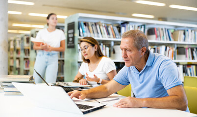 Wall Mural - Concentrated mature man spending time in library, reading books and making laptop. Self-education concept