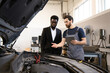 © sofiko14 - Mechanic in work overalls explaining car issues to customer in suit in modern auto repair shop. Professional consultation about vehicle maintenance and repair process.