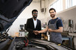 © sofiko14 - Mechanic and customer shaking hands in auto repair shop, sealing deal and expressing satisfaction. Mechanic holding tablet, customer dressed in business attire. Car hood open, tools visible.