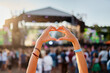 © artiemedvedev - Concertgoer forms heart shape with hands at beachside music festival, crowd enjoys live performance in sunset, love symbol gesture at summer event, youth celebration of art, outdoor entertainment.