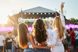 © artiemedvedev - Group of girls at beach music fest, arms raised, enjoying sunset. Friends hug, celebrate, dance in crowd, summer party vibe. Sunlight flares on stage, festival-goers gather, casual outdoor fun.