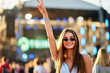 © artiemedvedev - Smiling young woman enjoys summer music festival, raises peace sign, wears sunglasses, wristband, against stage lights. Happy crowd, outdoor concert vibes, live performance event at beach.