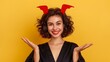 © Trushchenko_O - A woman with red horns on her head is smiling and waving. The image has a fun and playful mood. Devil girl isolated on yellow color background. Studio portrait of woman in spooky Halloween costume.