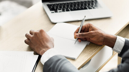 Leinwandbilder - A person's hand writes in a notebook while sitting at a desk, a laptop in the background.