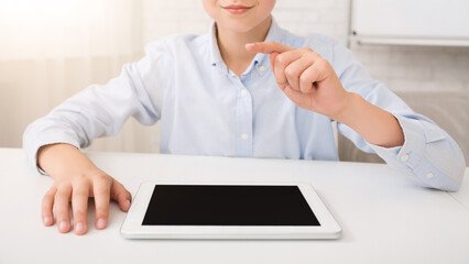 Poster - A young boy sits at a white desk in a bright room. He is wearing a light blue button-down shirt and is using a digital tablet with a black screen. He is pointing with his index finger at the tablet.