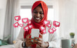 © Prostock-studio - African American woman wearing a red hijab smiles while looking at her smartphone, surrounded by pink heart icons representing social media likes. She is sitting at a table in a bright, modern room.