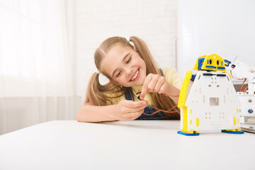 Poster - A young girl with pigtails smiles as she focuses on connecting wires to her robot. The robot is white and yellow, and the girl is sitting at a table in a brightly lit room.