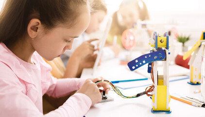 Aufkleber - A young girl in a pink shirt is focused on building a robot in a classroom setting. She is carefully connecting wires and parts with her small hands