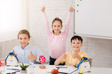 Poster - Three young students are sitting at a desk in a classroom setting, giving thumbs up and smiling. The girl in the back is wearing a pink shirt and has her arms raised in the air
