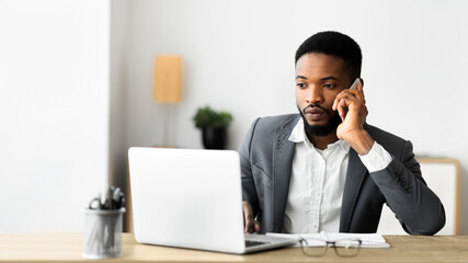 Poster - Focused african american businessman talking on cellphone and working on laptop in modern office, panorama with copy space