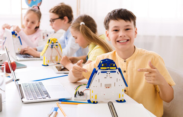 Aufkleber - A young boy sits in a classroom, beaming with pride as he points to a robot he built. He is surrounded by other students who are also engaged in STEM activities.
