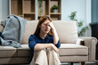 © Phushutter - A young Asian woman sits on a sofa in her living room, feeling sick with mental health diseases. She struggles with depression, anxiety, and other conditions, seeking support and solace at home.