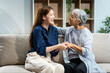 © Phushutter - A mature mom and a young Asian woman, mother and daughter, sit together on a sofa in their living room, celebrating Mother's Day with love and bonding at home.