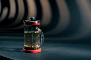 on a dark blue background with side lighting there is glass modern teapot with a press brewing green tea