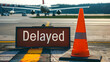 © sornram - Airport runway with delayed sign, traffic cone, and airplane in the background. Representing travel delay and transportation issue.