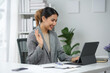 © amnaj - Professional woman conducting video conference at home office, working remotely, waving and smiling at laptop during virtual meeting.