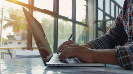 Sticker - Hands type on a laptop as morning sunlight filters through large windows, creating a warm, productive work atmosphere.